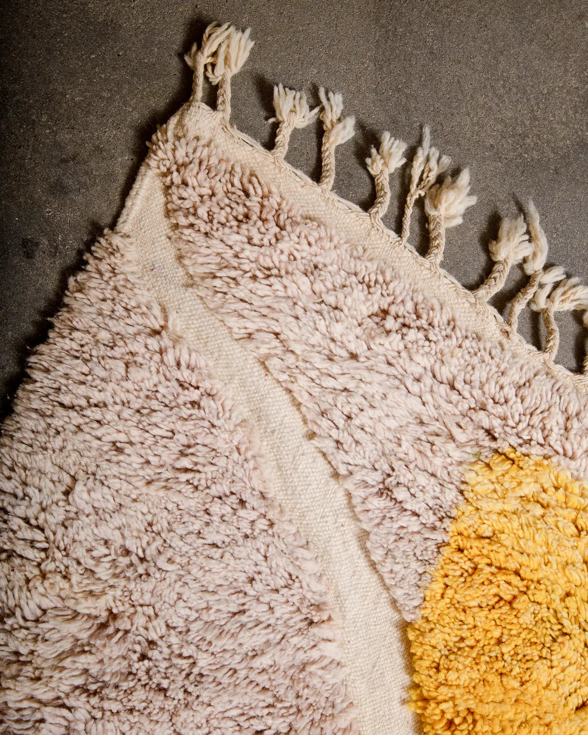 Tasseled edge of vintage Berber rug featuring fluffy wool and contrasting flat woven design laying on concrete studio floor.